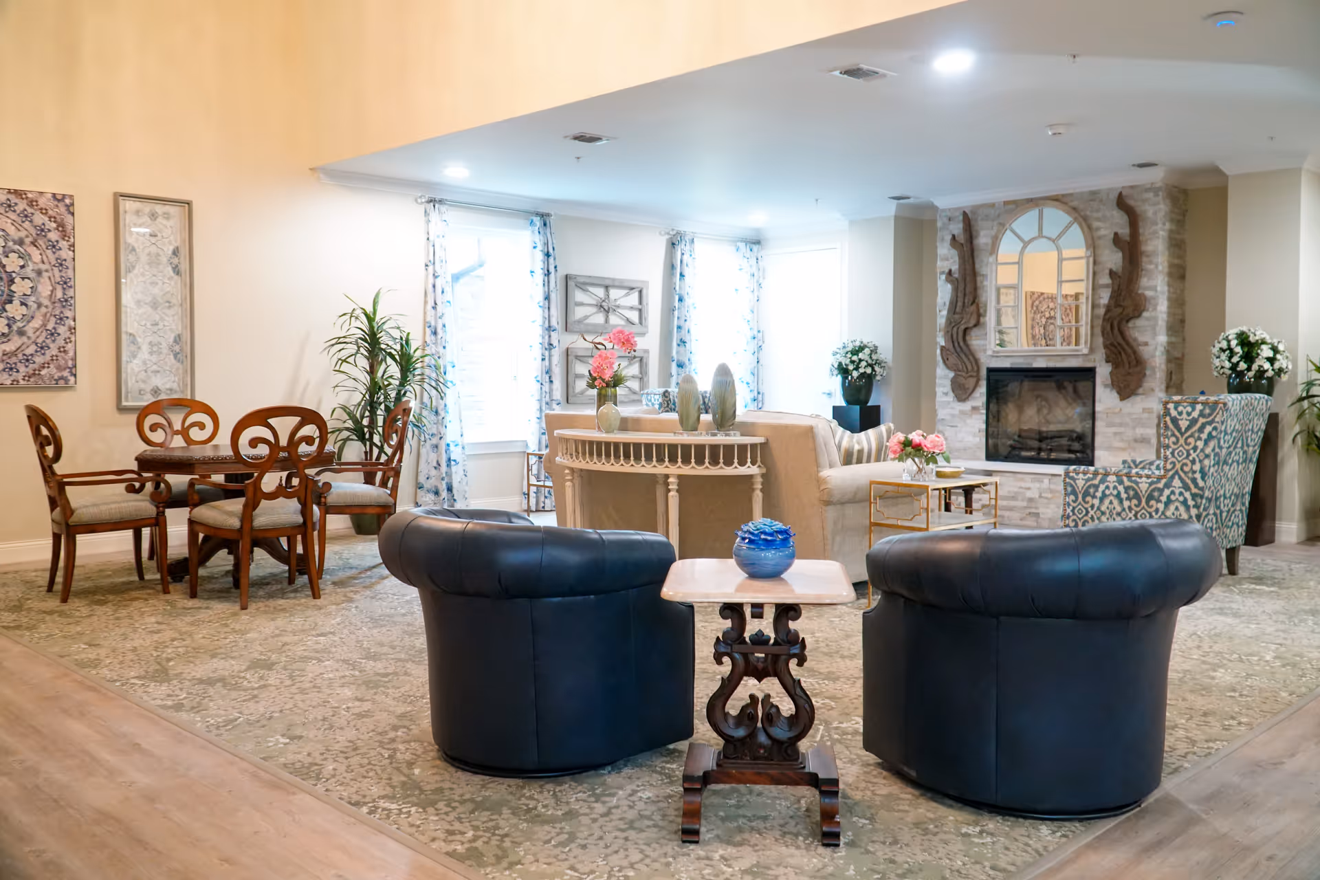 A cozy living room area with two black leather chairs facing a small wooden table with a blue decorative item. Behind them is a beige sofa with a console table adorned with vases and flowers. To the right, there is a patterned armchair next to a stone fireplace with a large mirror and decorative wooden wall hangings. On the left side, there is a wooden dining table with four chairs and framed artwork on the wall. The room has light-colored walls, large windows with floral curtains, and a patterned carpet.