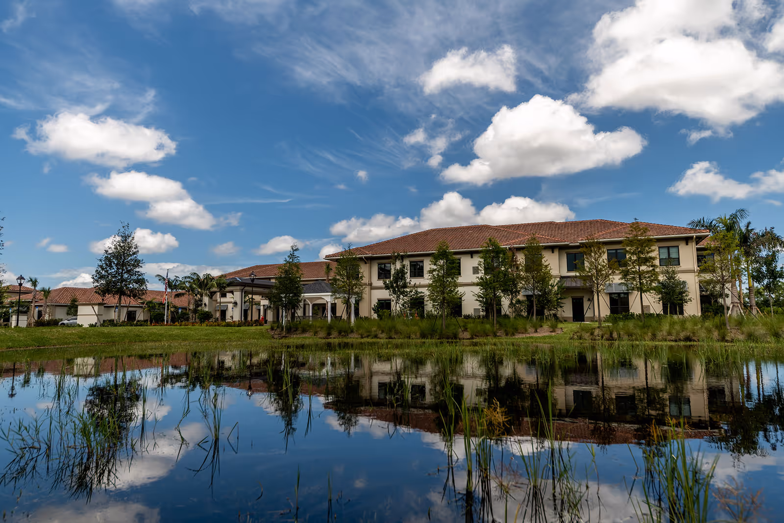 Exterior view of The Capstone at Royal Palm Senior Living building with a red-tiled roof, surrounded by trees and greenery, reflected in a calm pond under a blue sky with scattered clouds.