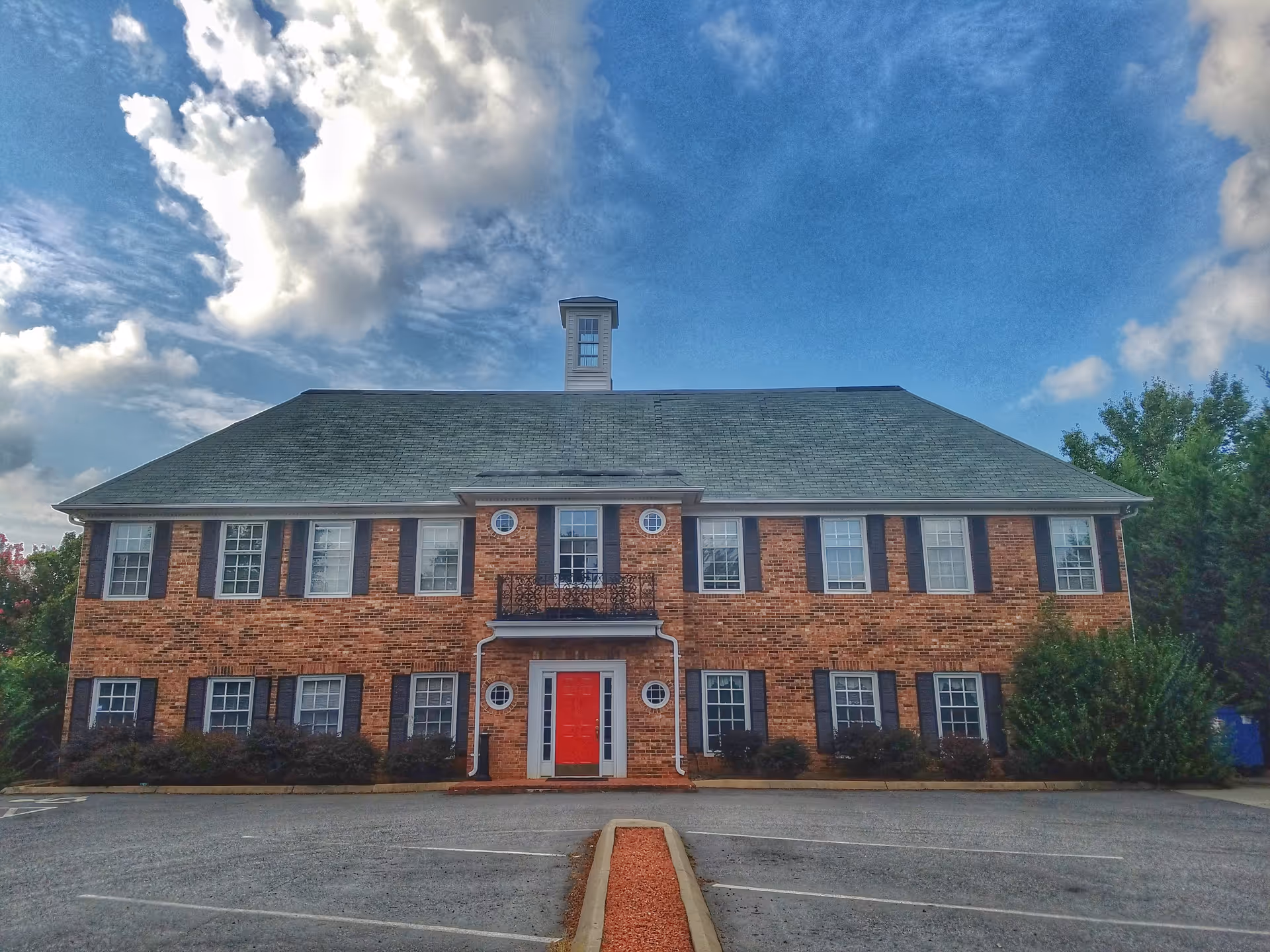 Brick two-story building with red front entrance and surrounding shrubs under blue sky.