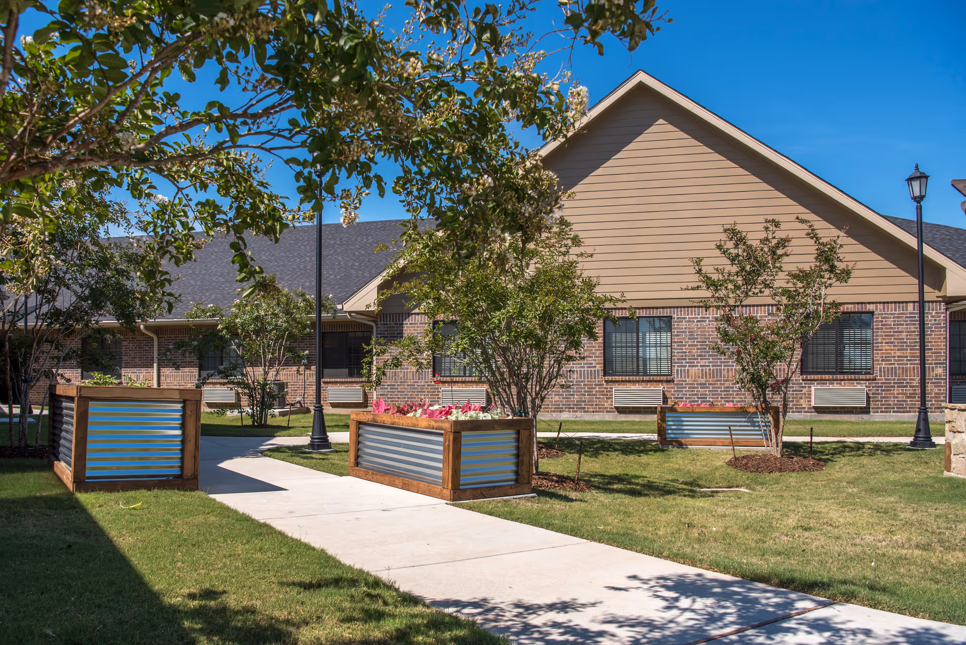 Single-story brick and siding senior living building with a paved walkway, raised planters, trees, and lampposts in a sunny courtyard.