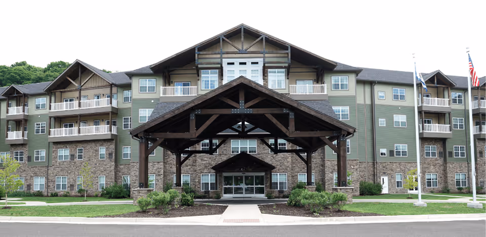 Front exterior view of a large senior living facility building with multiple floors, balconies, and a prominent covered entrance supported by wooden beams. The building features a combination of stone and green siding with several windows and flagpoles with flags on the right side.