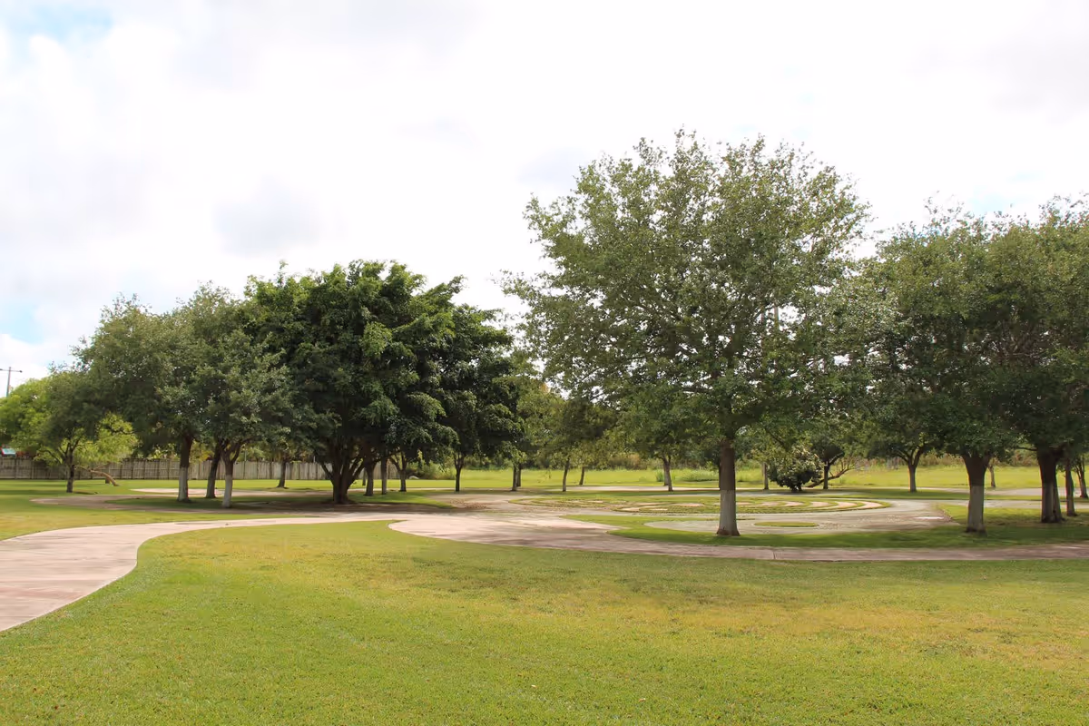 Grassy park-like lawn with a winding paved path and several trees under a partly cloudy sky.