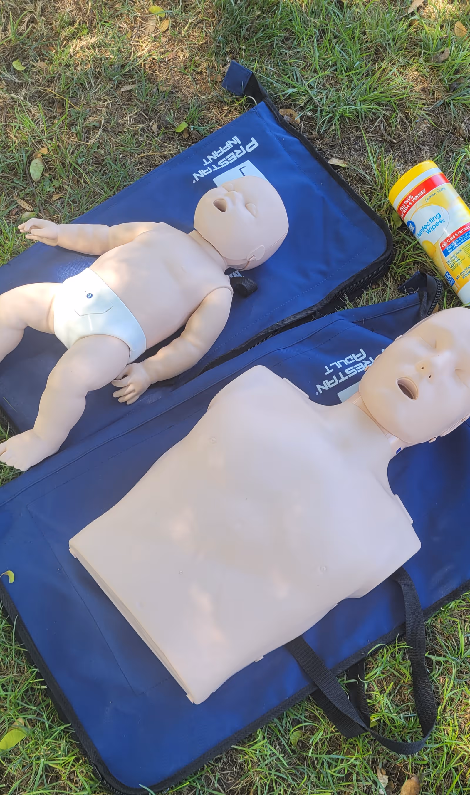 Two CPR mannequins, adult and infant, lie on blue mats outdoors on grass.
