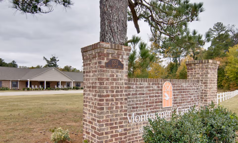 Brick entrance sign reading 'Montgomery Gardens' with a small address plaque on a pillar and the single-story facility building and grounds visible behind it.