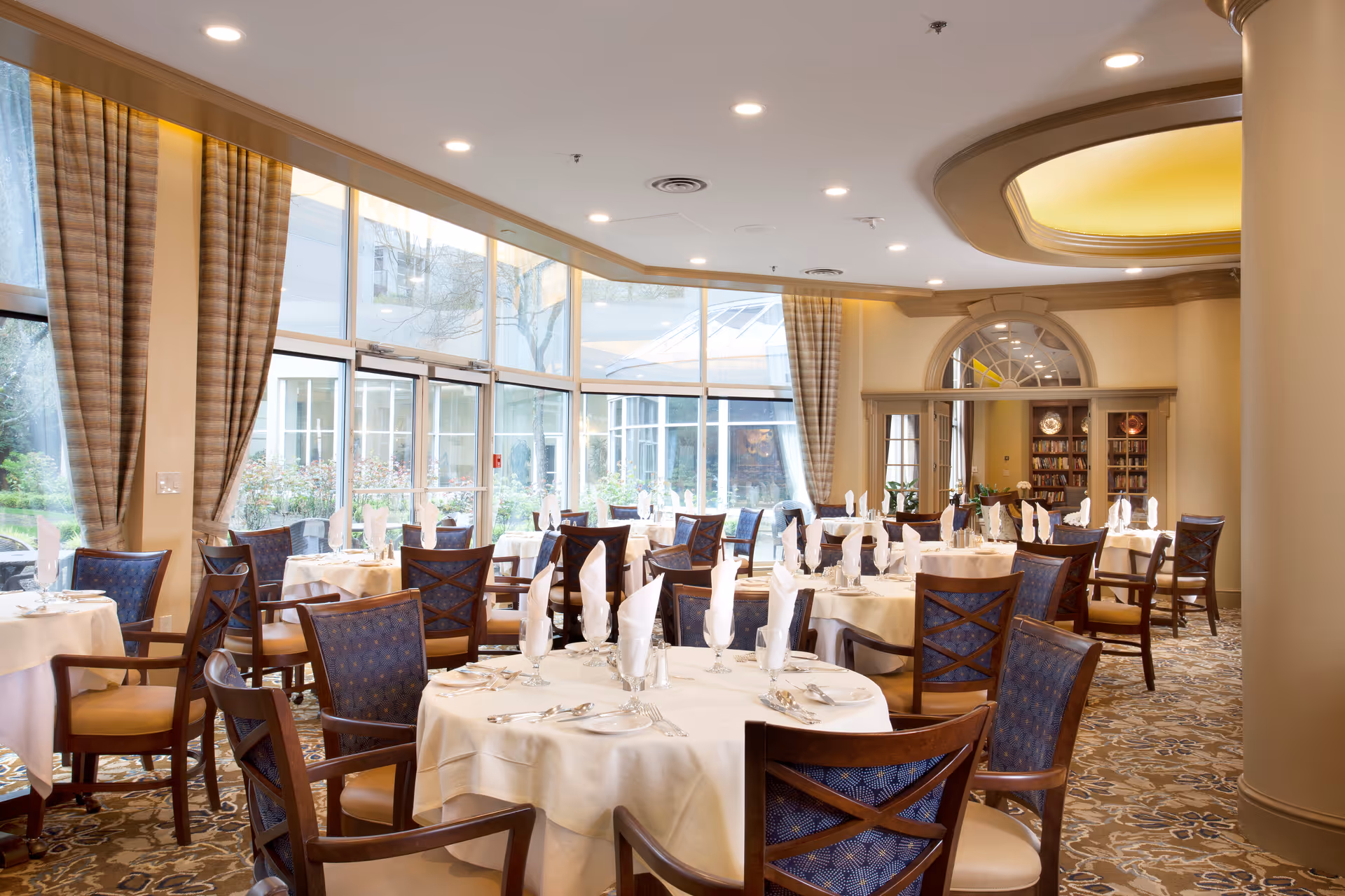 Elegant dining room with round tables, white linens, and tall windows in a senior living facility.