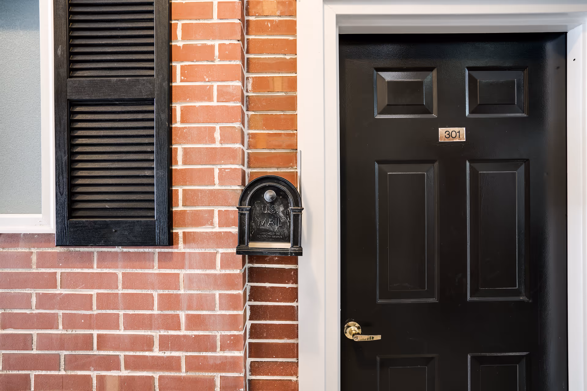 Brick exterior with a black front door, mailbox, and window shutters.