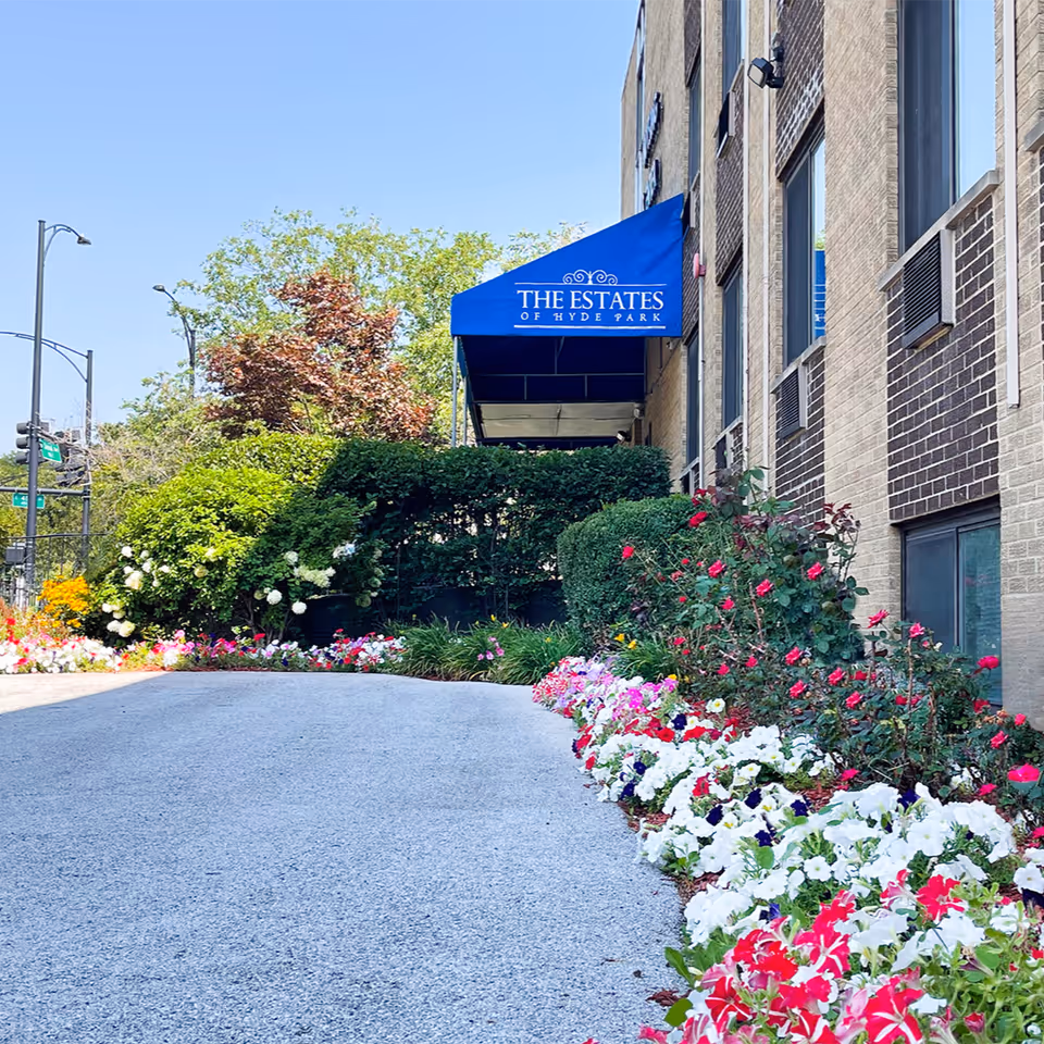 Driveway bordered by colorful flower beds leading to a brick building entrance with a blue canopy reading "THE ESTATES OF HYDE PARK".