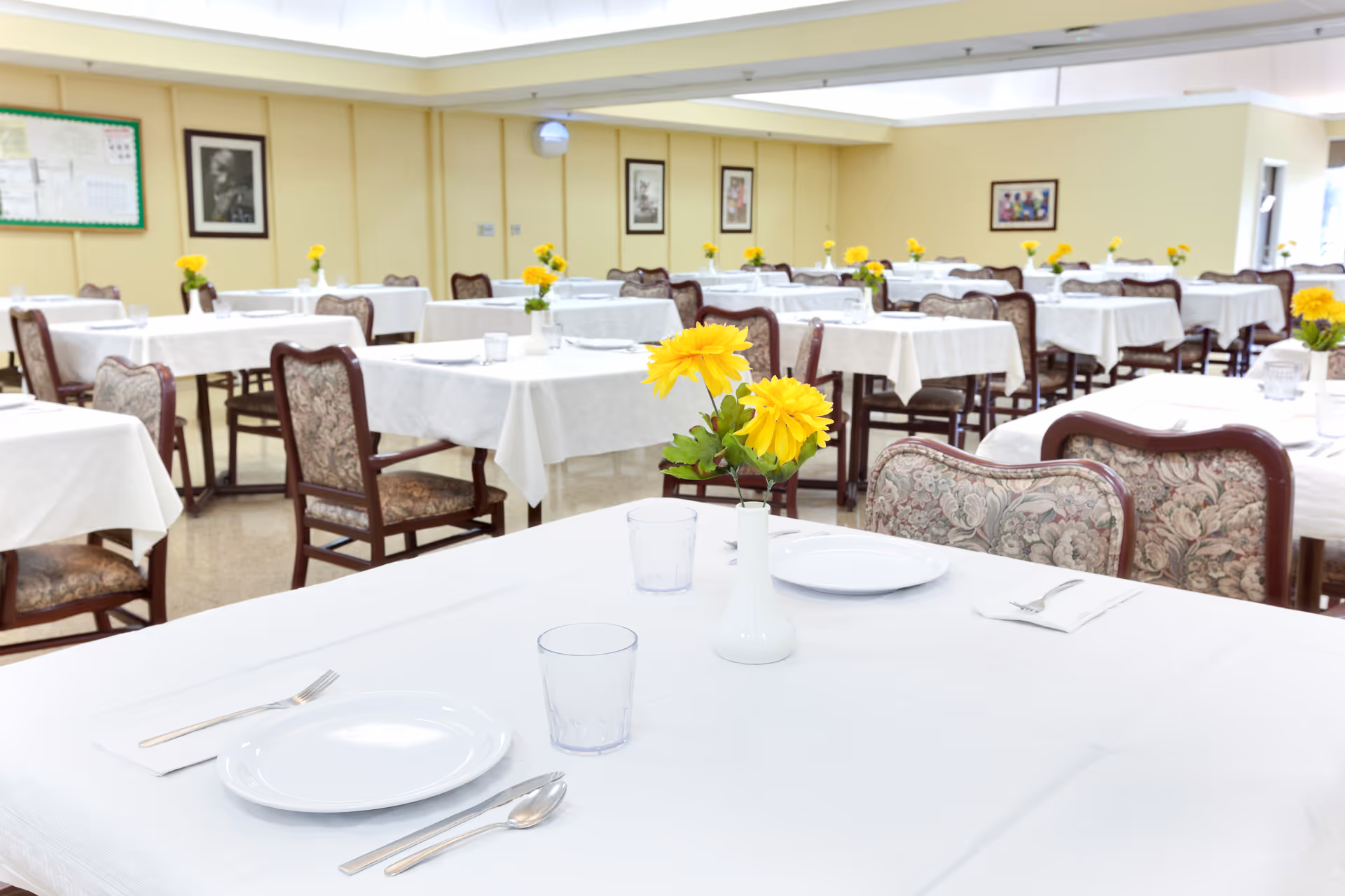 Bright dining hall with tables set for meals and yellow flowers.