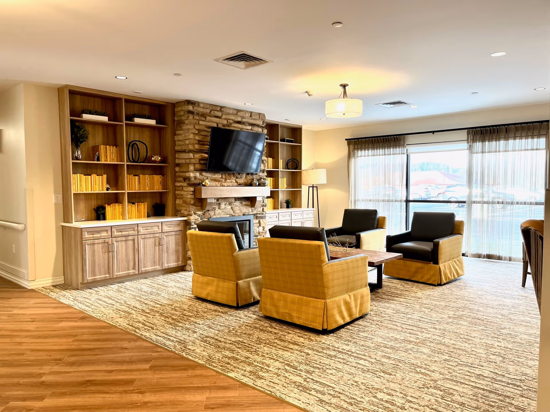 Cozy communal living room with yellow chairs, wood shelves, a stone fireplace, and large windows.