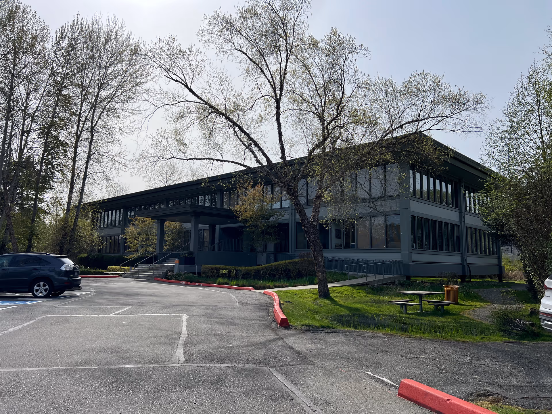 Two-story modern building with many windows, parking lot, trees, and an outdoor picnic table.