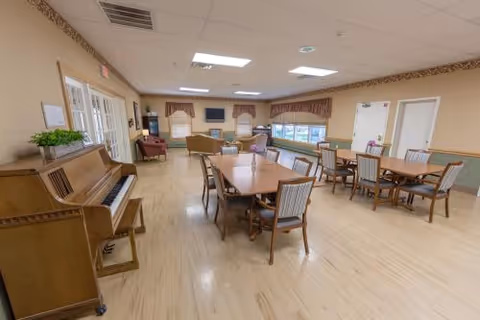 A spacious common area with several wooden tables and chairs arranged neatly on a light wood floor. There is a piano on the left side with a small plant on top, and comfortable seating including armchairs and sofas near the windows at the far end. The walls are beige with a decorative border near the ceiling, and there are windows with valances letting in natural light. The ceiling has recessed lighting and a few ceiling tiles.