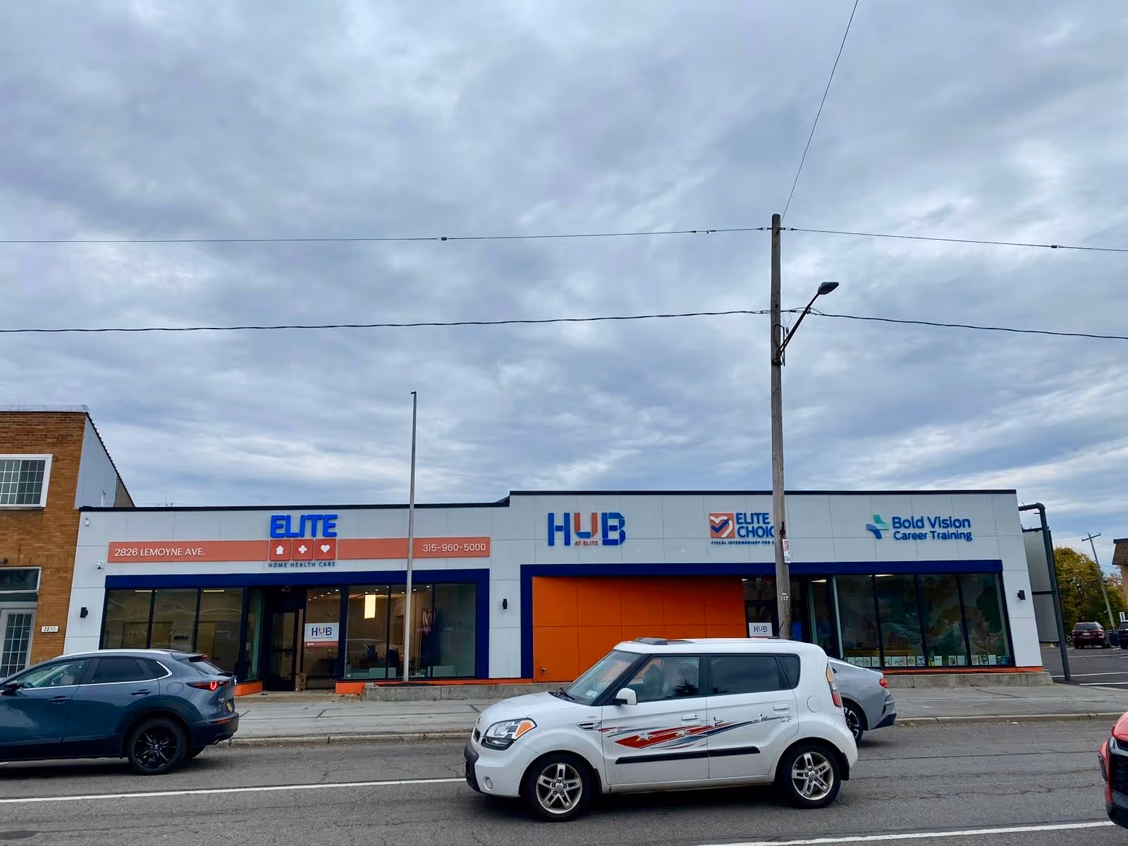 Exterior view of a storefront strip with Elite and Hub signs and parked cars