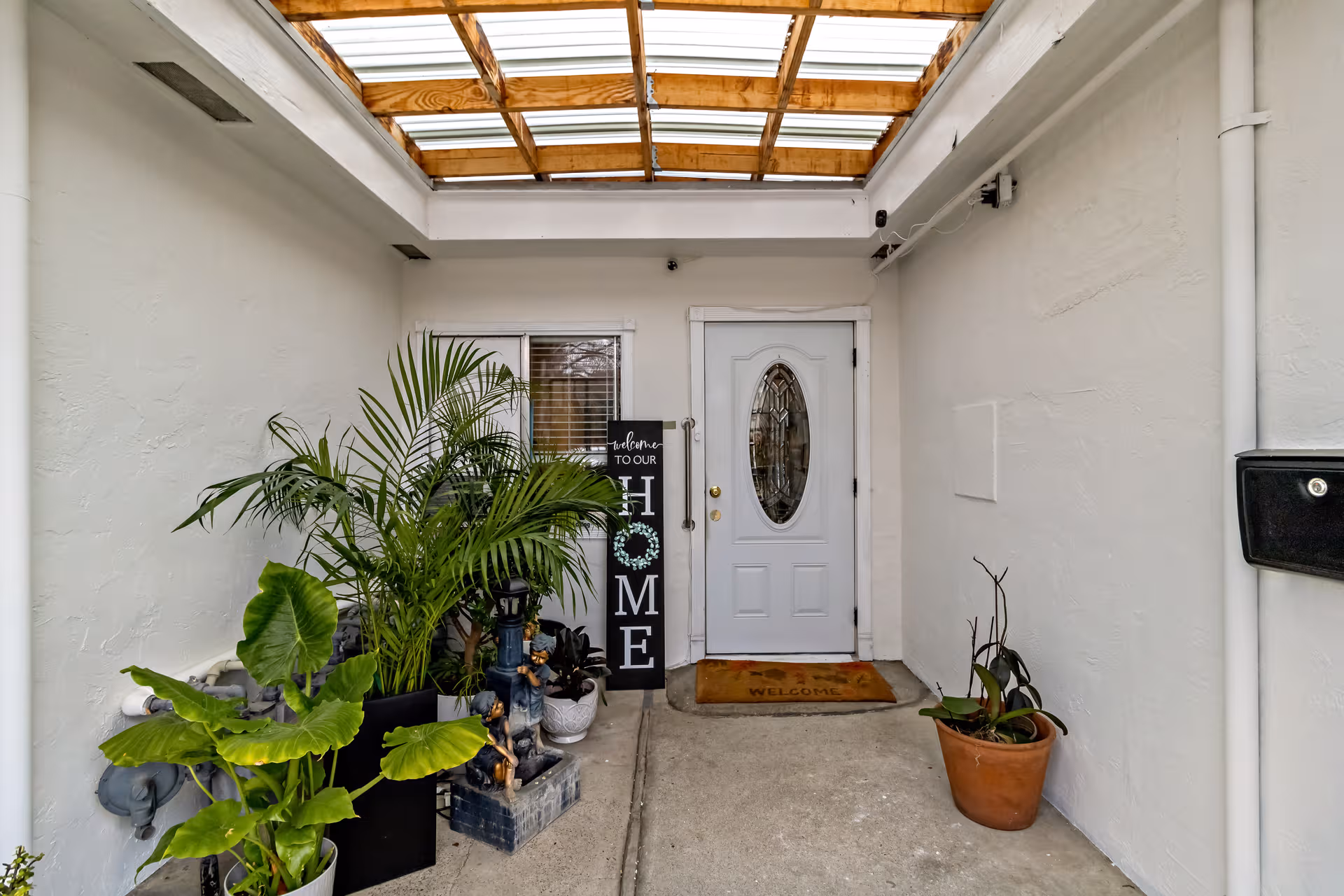 A small outdoor entry with potted plants and a front door under a skylit covered walkway.