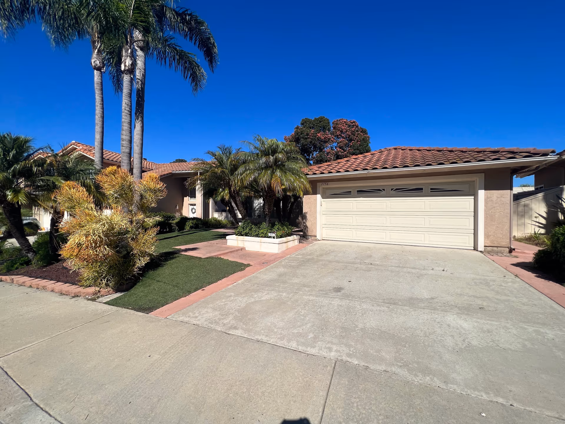 Single-story Mediterranean-style house with a tiled roof, two-car garage, palm trees, and a driveway under a clear blue sky.