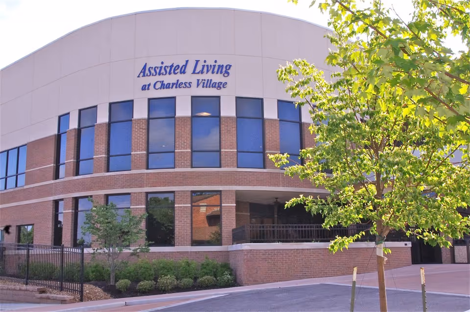 Exterior view of a two-story assisted living facility building with large windows and a sign that reads 'Assisted Living at Charless Village'. There are trees and shrubs in front of the building and a paved driveway or parking area.