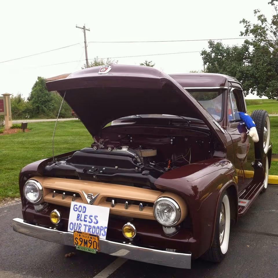 Maroon vintage car with hood open parked outdoors; sign reads God Bless Our Troops.