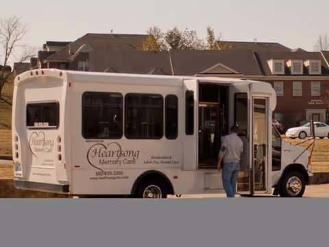 White Heartsong Memory Care shuttle bus parked outside a residential facility, with a person boarding.