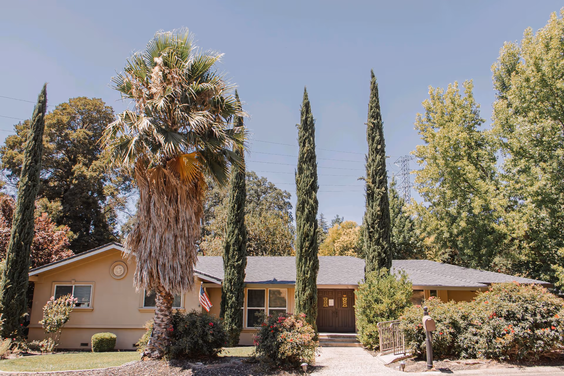 Single-story beige senior living building with tall trees and a landscaped front yard.