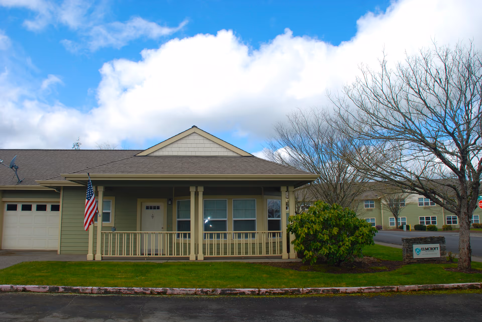 Exterior view of a single-story residential building with a front porch, an American flag hanging on a post, a garage door on the left, and a green lawn with a bush and a tree. The sky is partly cloudy with blue patches. A sign near the tree reads 'ELMCROFT OF STAYTON'.