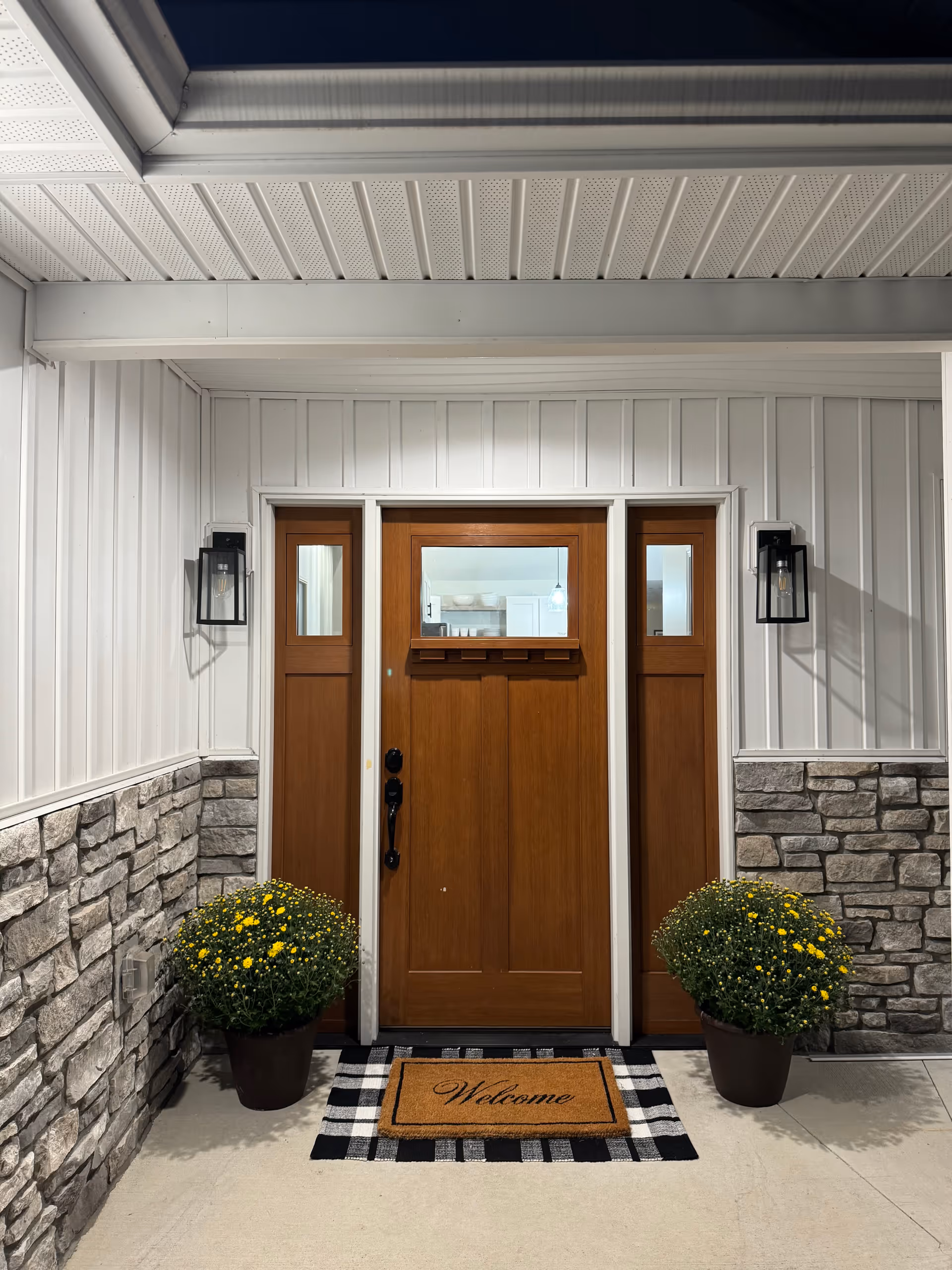 Front entrance of a senior living facility with wooden door, stone walls, planters, and welcome mat.