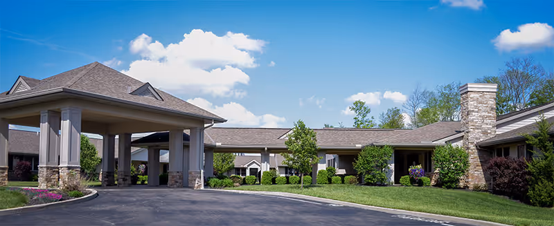 Single-story senior living building with a covered porte-cochère entrance, stone columns, and landscaped lawn under a blue sky.