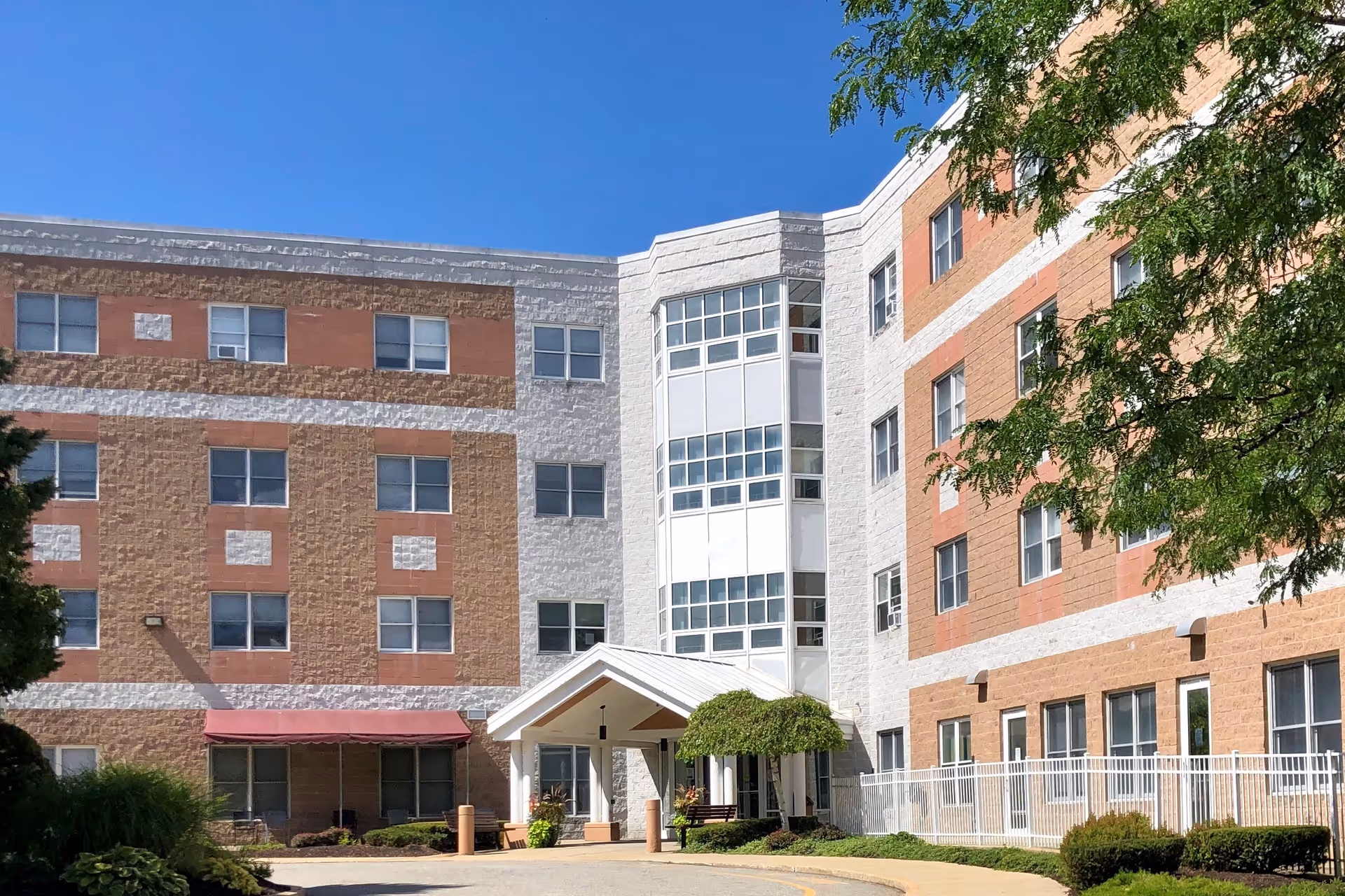 Exterior view of Oakland Grove Health Care Center showing a multi-story building with a mix of beige, white, and reddish-brown brickwork. The entrance has a white covered portico with columns and is surrounded by shrubs and small trees. The sky is clear and blue.