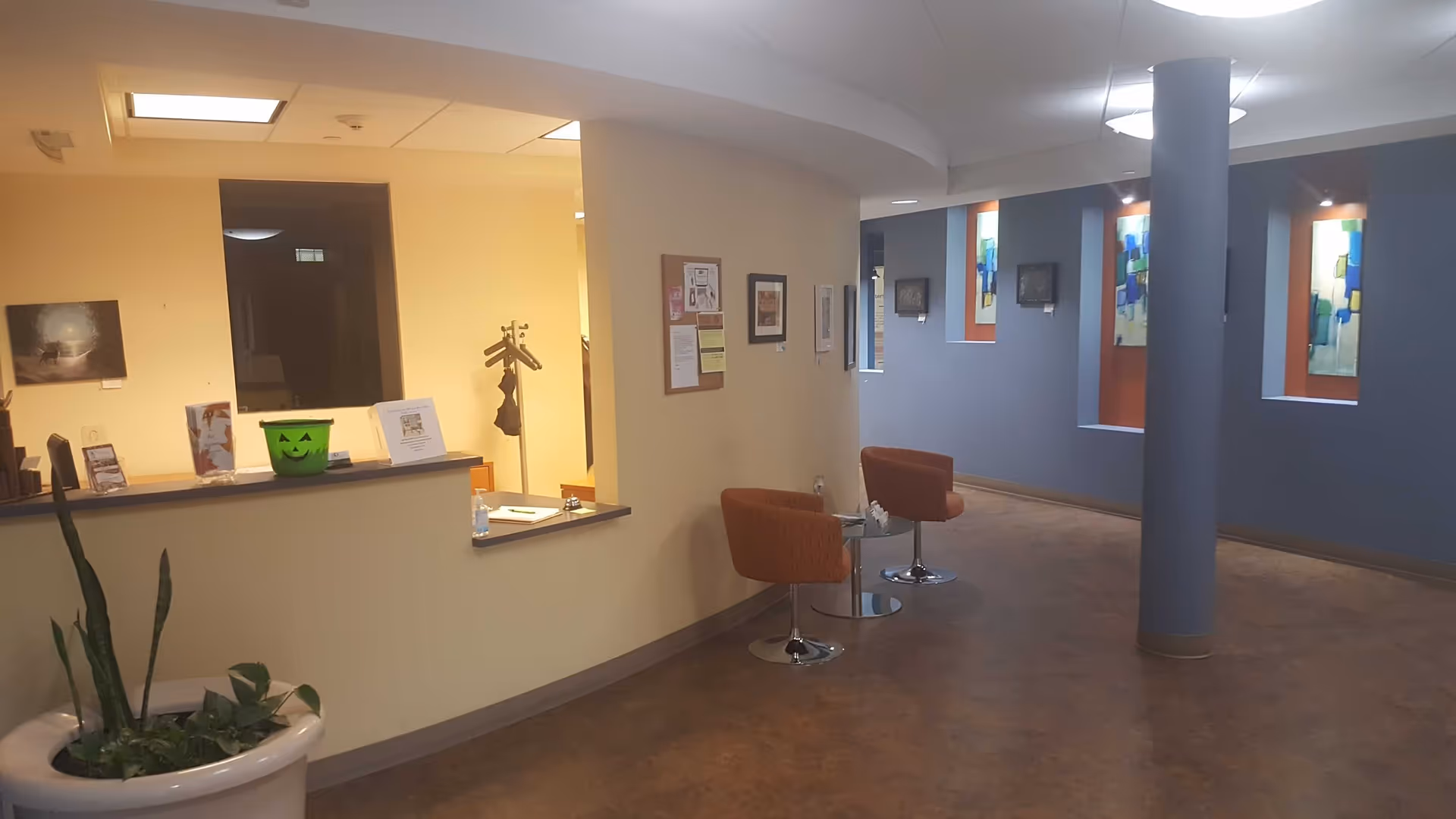 Reception area with beige walls, curved desk, orange chairs, and blue wall art.
