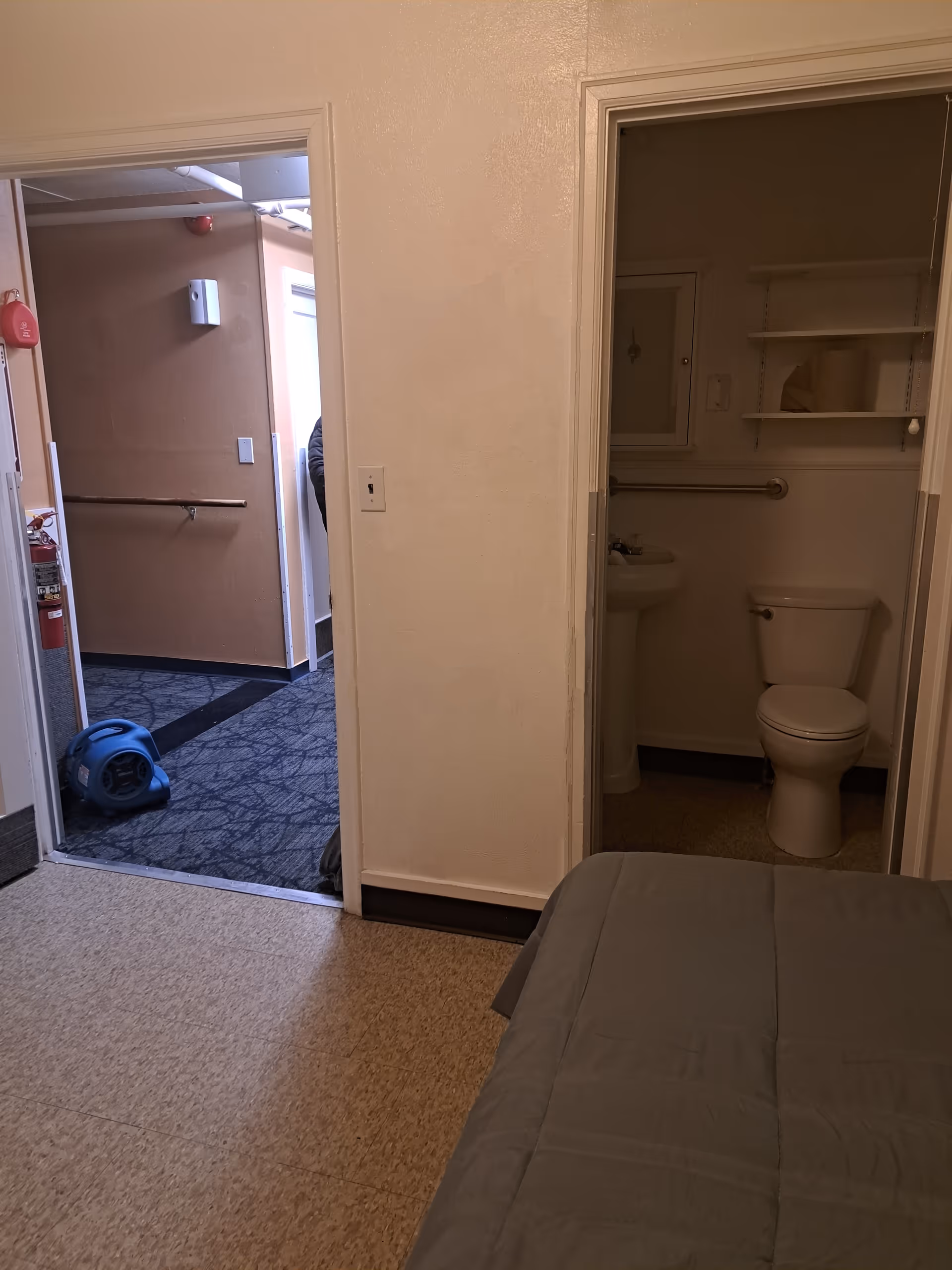 View from a bedroom showing part of a bed with a gray blanket, an open doorway leading to a small bathroom with a toilet and sink, and another open doorway leading to a hallway with a blue floor fan and a fire extinguisher mounted on the wall.