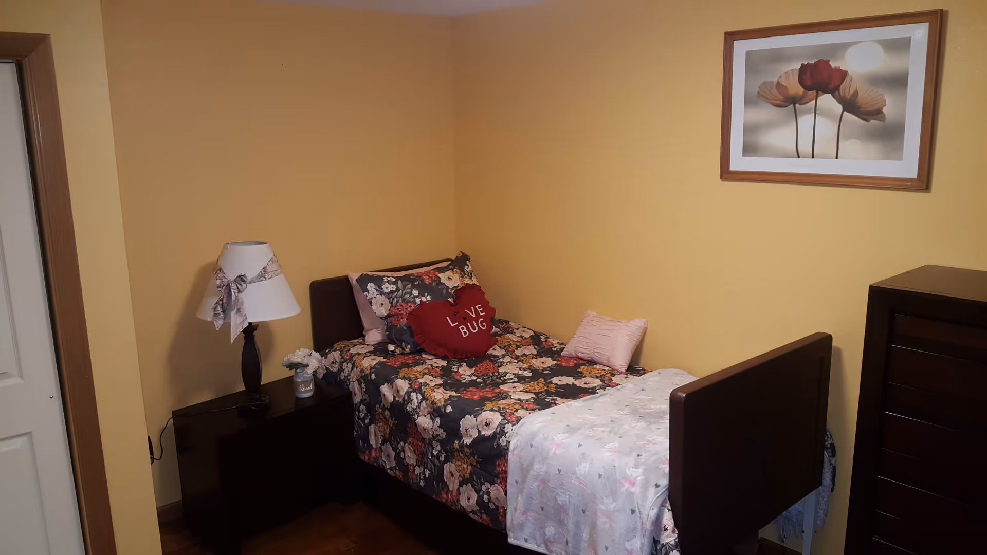Small yellow-walled bedroom with a floral bed, nightstand lamp, dresser, and framed flower art.