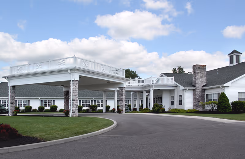 Exterior view of a single-story white building with a covered entrance supported by stone pillars, surrounded by green lawns and shrubs under a partly cloudy blue sky.