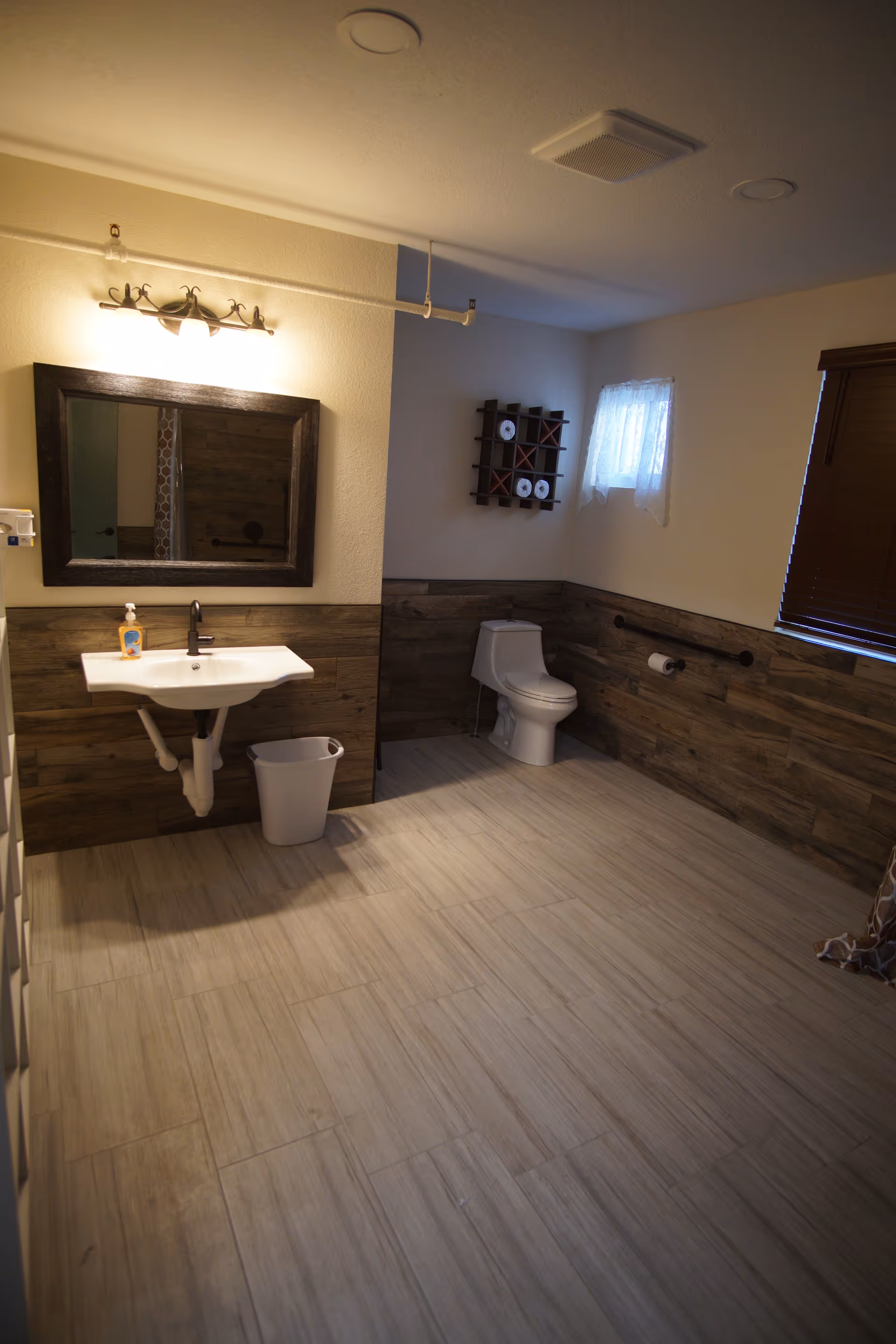 A spacious bathroom with light-colored tiled floor and wood-paneled lower walls. The room features a white toilet, a white wall-mounted sink with a soap dispenser, a large rectangular mirror above the sink, a small window with a sheer curtain, and a wall-mounted rack holding toilet paper rolls. There are grab bars installed near the toilet and a trash bin under the sink.