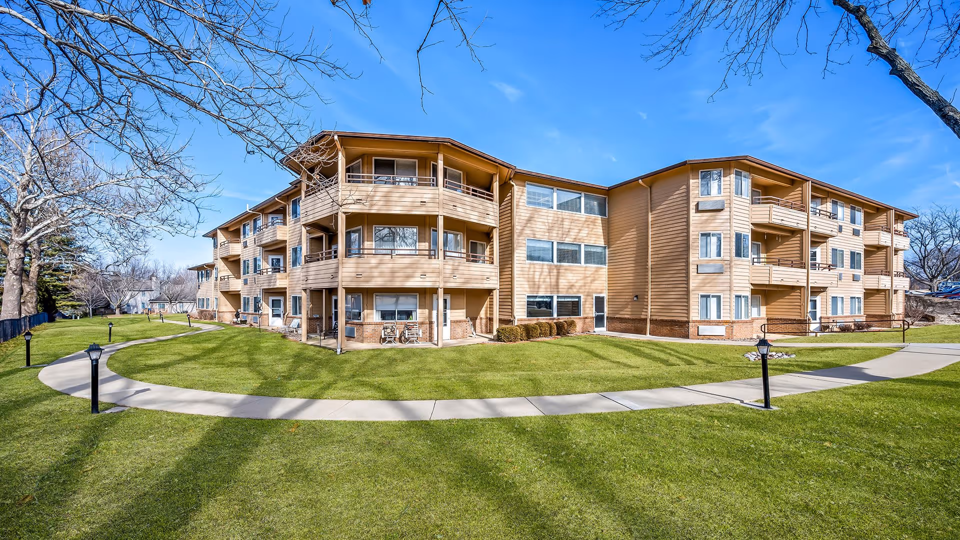 Exterior view of a three-story senior living facility building named Holiday Blair House with beige siding and multiple balconies. The building is surrounded by a well-maintained green lawn, a curved concrete walkway, and leafless trees under a clear blue sky.