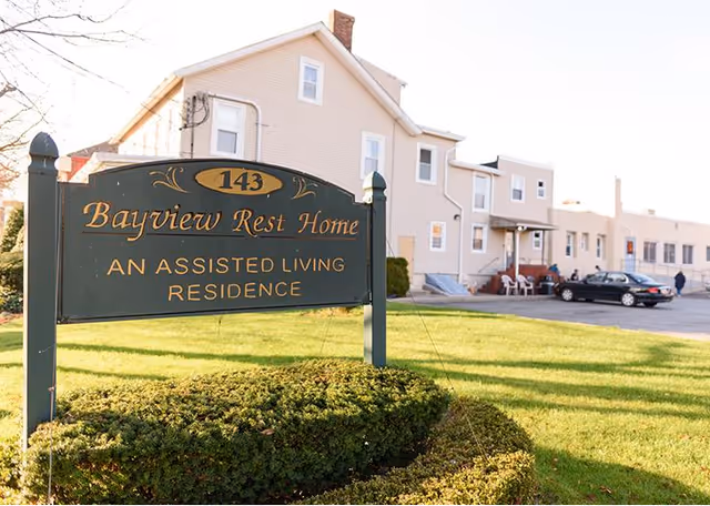 A green and gold sign reading '143 Bayview Rest Home An Assisted Living Residence' is displayed in front of a beige multi-story building with several windows and a parking area with cars. The scene is set on a sunny day with green grass and bushes around the sign.