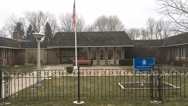 Single-story hospital entrance with flagpole, bench, and a manicured front lawn.