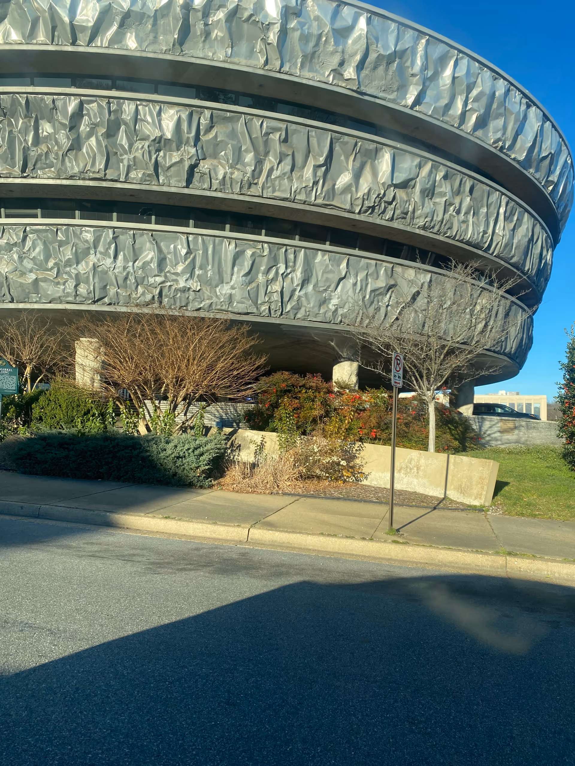Exterior view of a curved, metallic-clad building with landscaped bushes along the sidewalk.