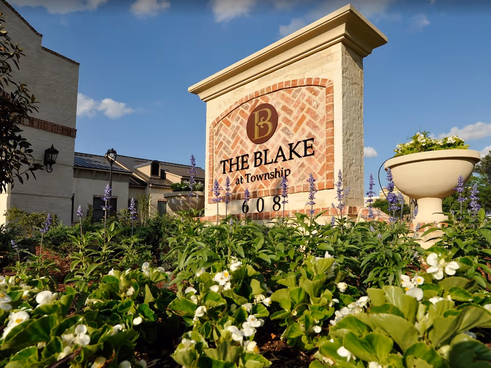 Outdoor view of a brick and stone sign for The Blake at Township surrounded by green plants and flowers with a building and blue sky in the background.