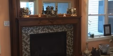 A cozy living room area featuring a wooden fireplace mantel with decorative items on top, including small figurines and plants. Above the mantel is a mirror reflecting part of the room. To the right, there is a window with white blinds and a small table with decorative objects, including a cat figurine.