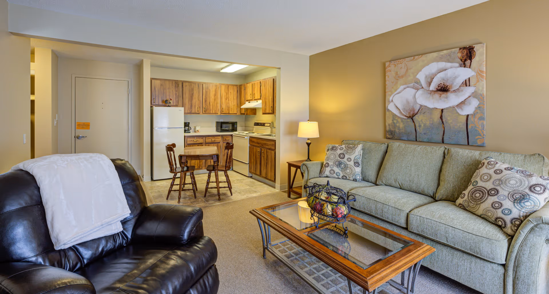 A cozy living area in a senior living community featuring a green upholstered sofa with patterned cushions, a glass-top wooden coffee table with a decorative fruit basket, a black leather armchair with a white throw blanket, a side table with a lamp, and a large floral painting on the wall. In the background, there is a small kitchen with wooden cabinets, a white refrigerator, stove, microwave, and a small wooden dining table with two chairs.