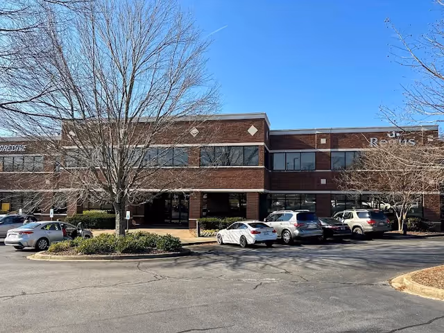 Two-story brick senior care facility entrance with a parking lot and leafless trees.