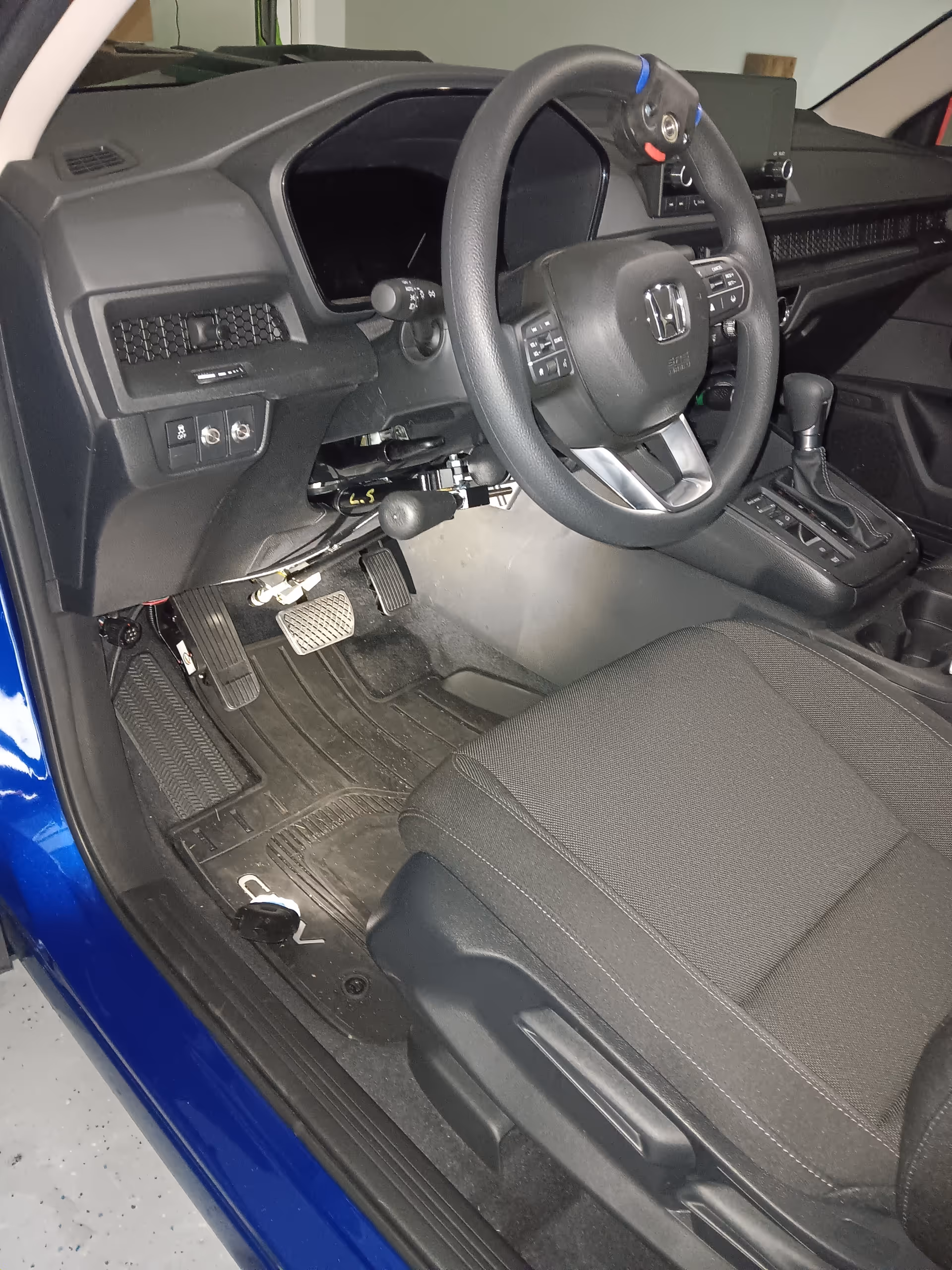 In-car view of a blue vehicle's steering wheel, dashboard, pedals, and front seat.