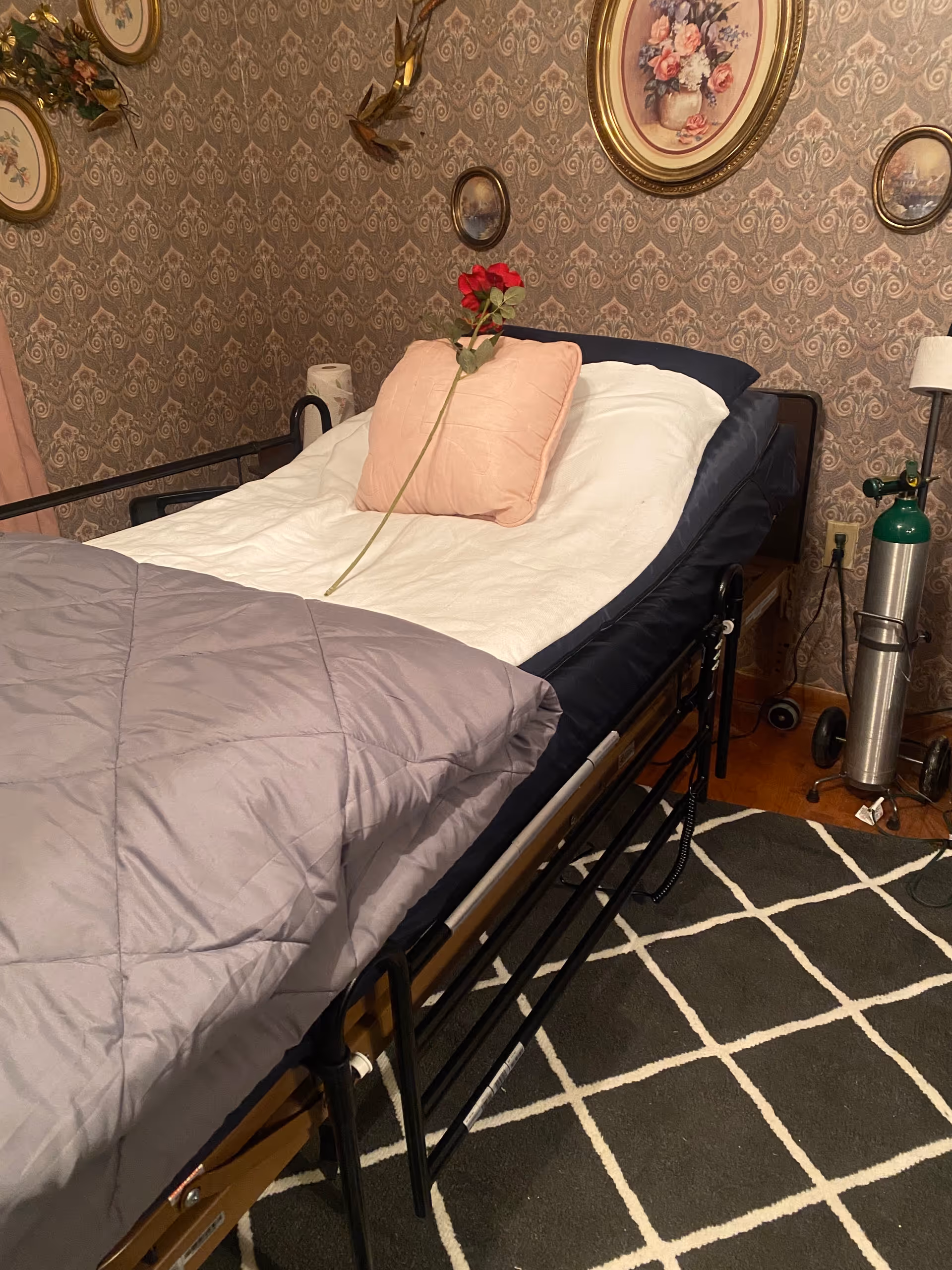 A hospital-style bed in a vintage-patterned bedroom, rose on pillow and medical oxygen tank nearby.