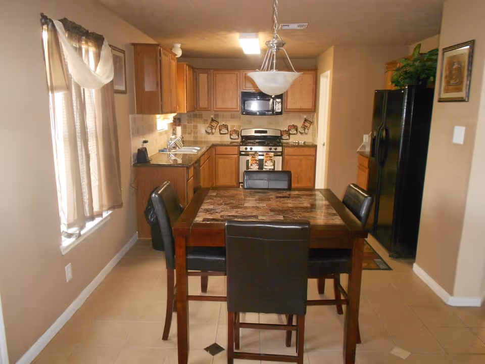 Dining table with four chairs in front of a galley kitchen with wooden cabinets, a stove and a black refrigerator.