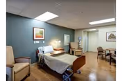 A patient room with a bed, seating, and warm lighting in a care facility.