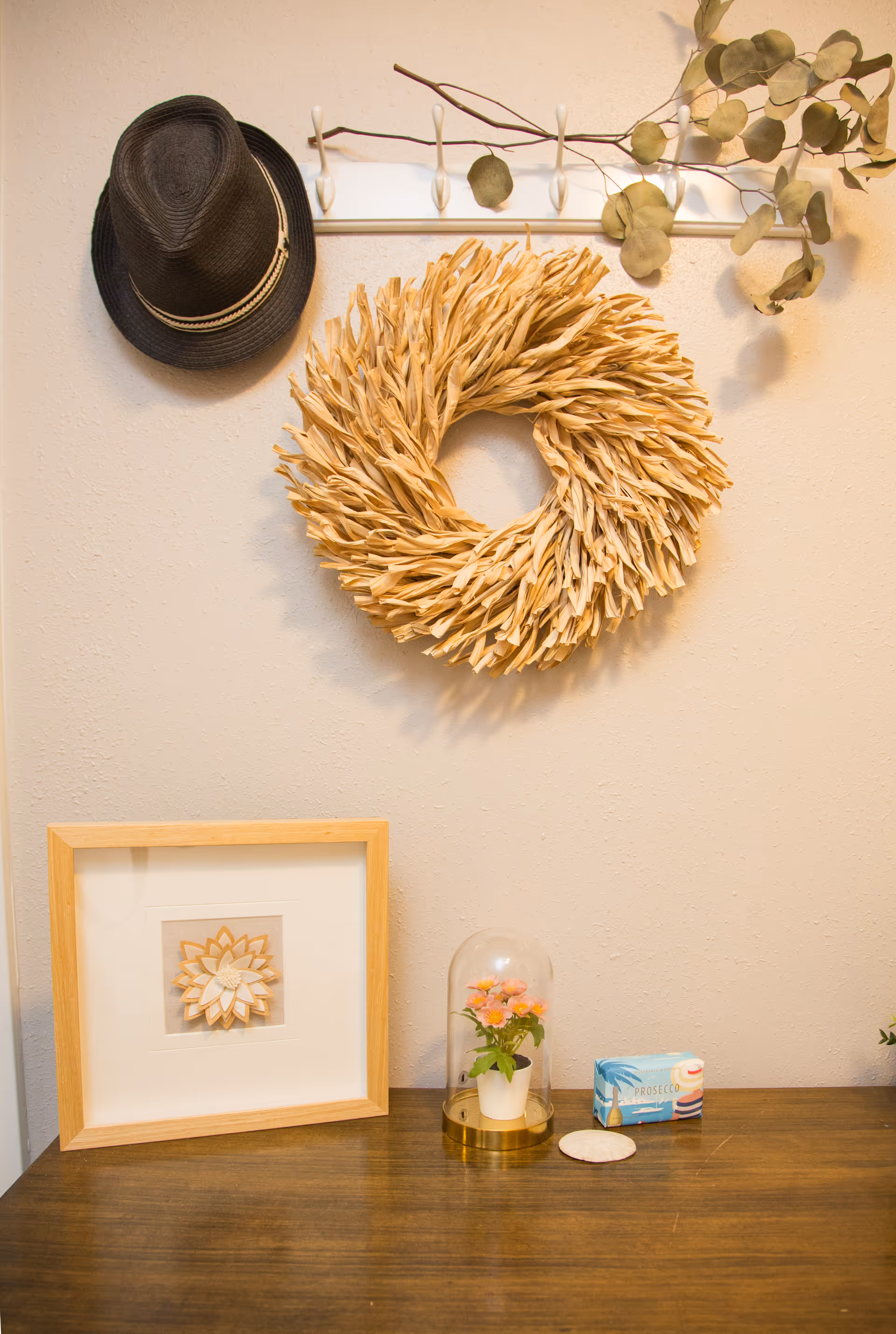 Decorative wall featuring wreath, hat, eucalyptus, framed art, and a glass cloche on a wooden table.