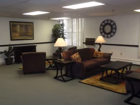 A communal lounge with a sofa, armchairs, side tables and lamps, a large wall clock, framed art, and potted plants.