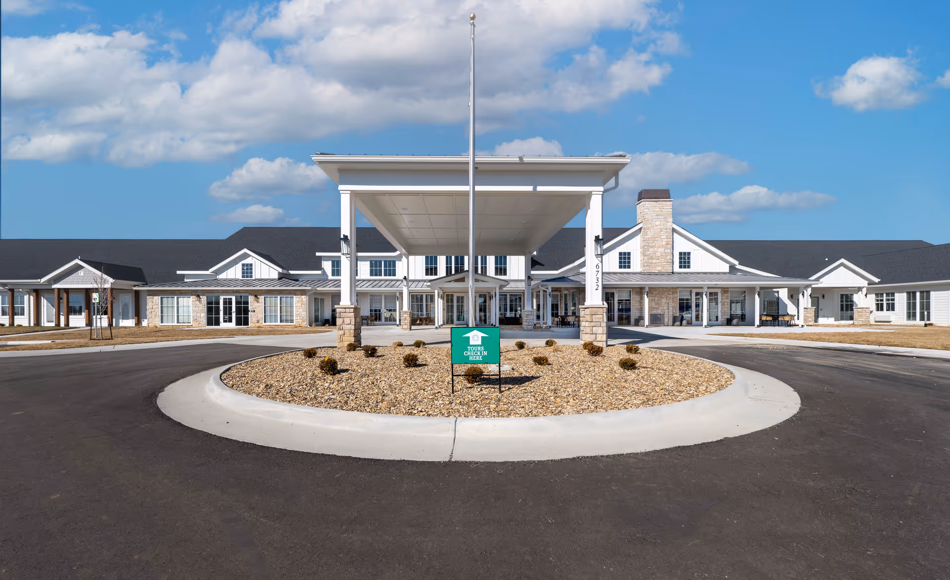 Front exterior view of Cedarhurst Senior Living of Topeka building with a covered entrance, a flagpole, and a circular driveway surrounded by landscaping under a partly cloudy blue sky.
