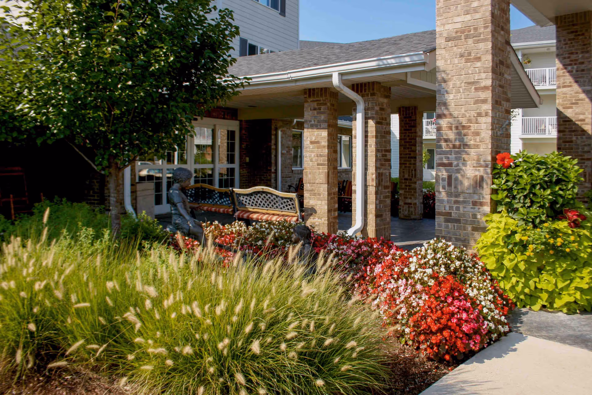 Covered brick entrance of a senior living facility with benches, a small statue, and colorful flower beds.