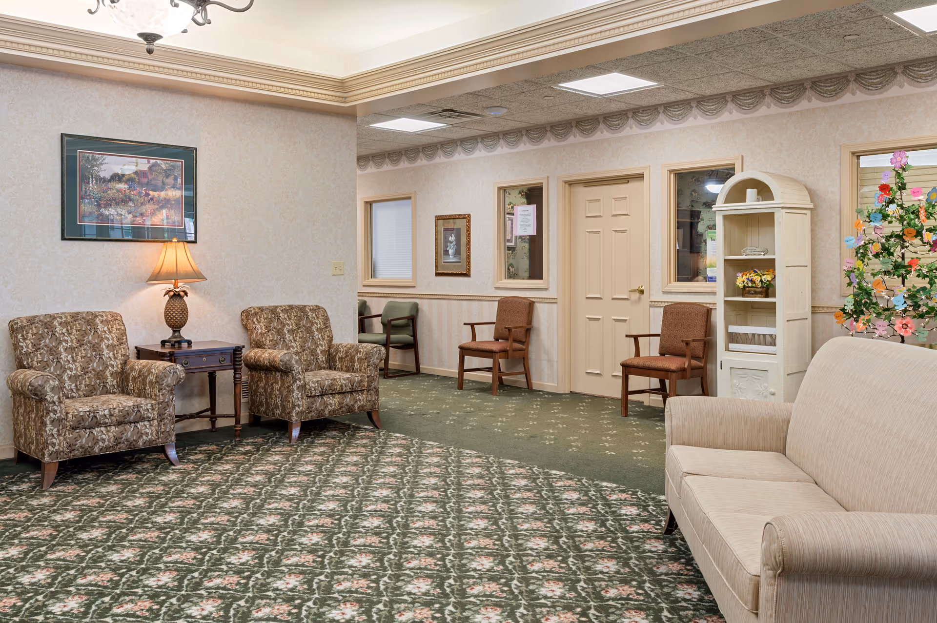 Interior lounge of a senior living facility with upholstered armchairs, a sofa, side table, lamp, and decorative shelving.