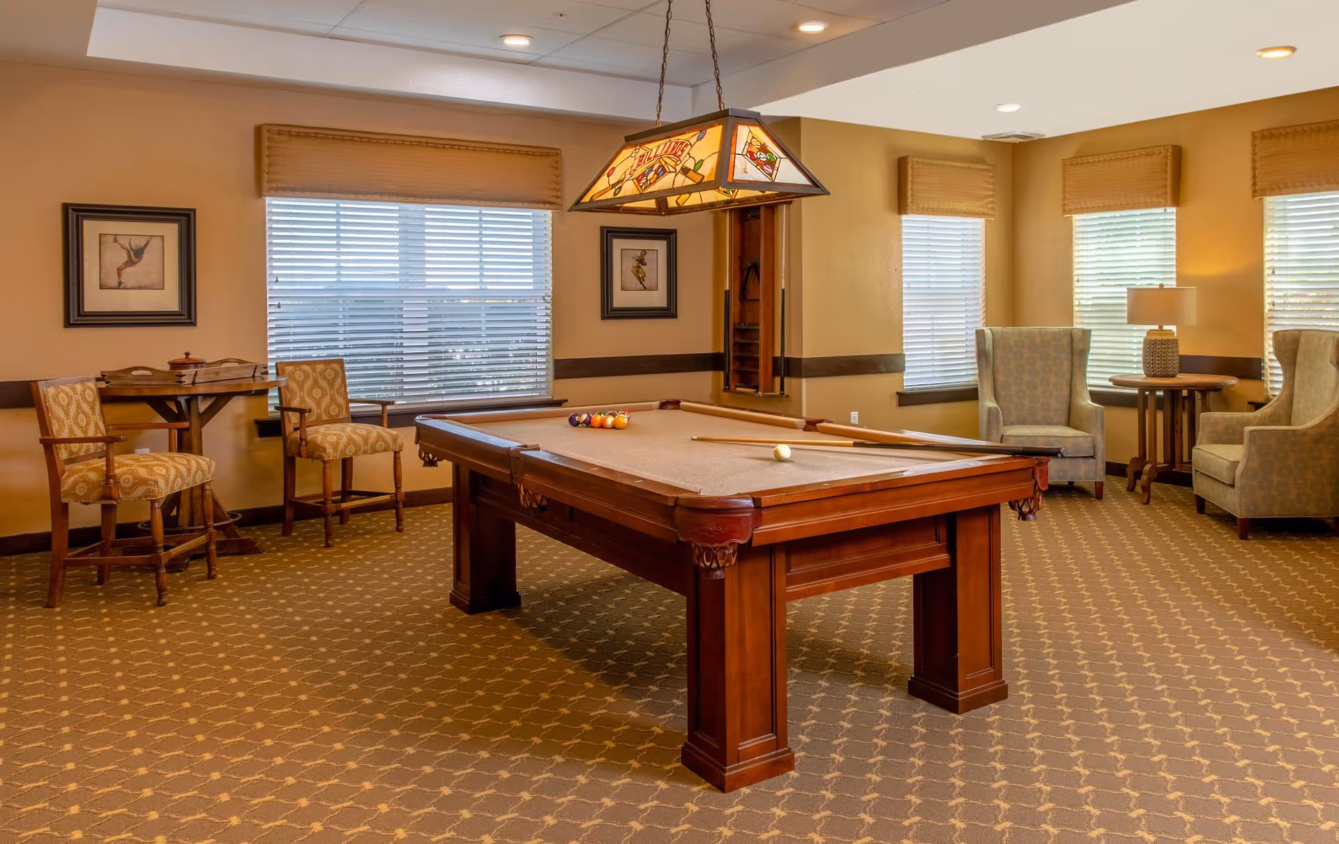 A well-lit recreational room with a pool table in the center, surrounded by comfortable chairs and a small table with two chairs near a window. The room has beige walls, patterned carpet, framed artwork, and a stained glass hanging light fixture above the pool table.