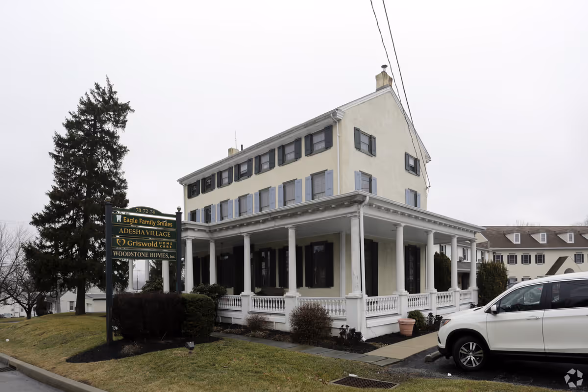 Beige multi-story senior living building with a white wraparound porch and front sign.